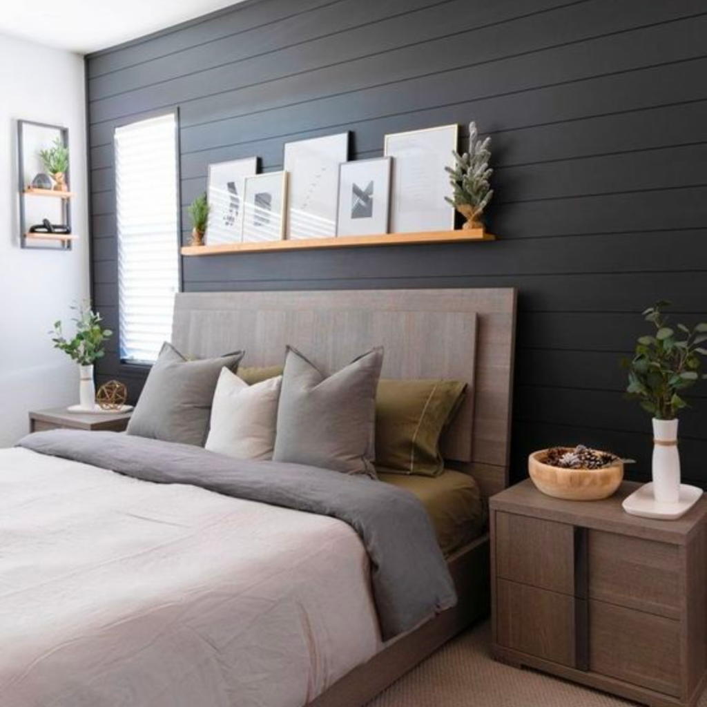 Farmhouse design bedroom with a black accent wall, a queen sized bed, medium tone nightstands over a neutral area rug.  Above the bed is shelving with artwork.
