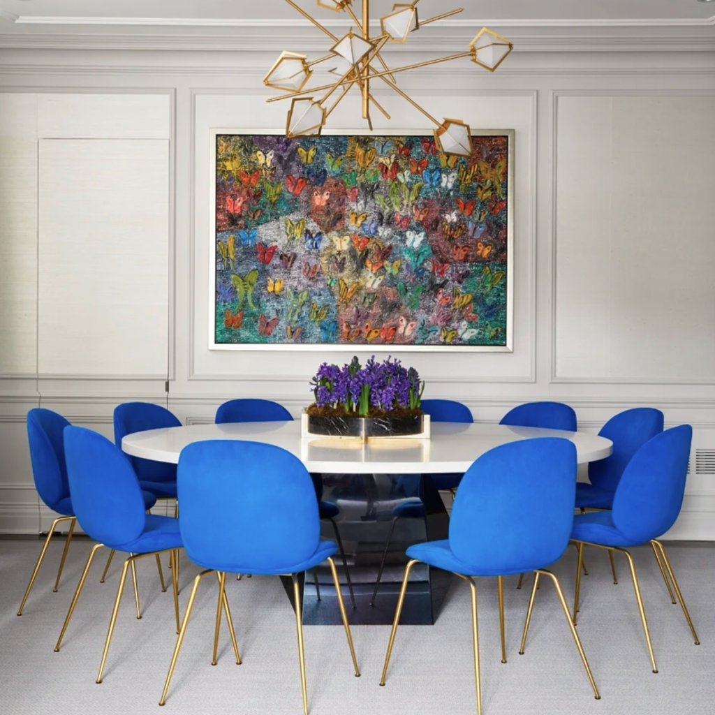 Colorful dining room with a round table and blue dining chairs.  Large, brass light fixture hung above the dining table.  Walls are built with custom trim by a skilled contractor.